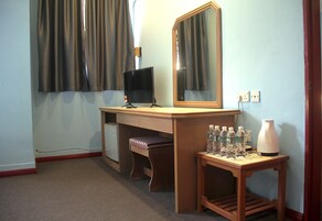 Habitación de hotel con escritorio, televisor, espejo, banco y pequeña mesa con botellas y un florero.