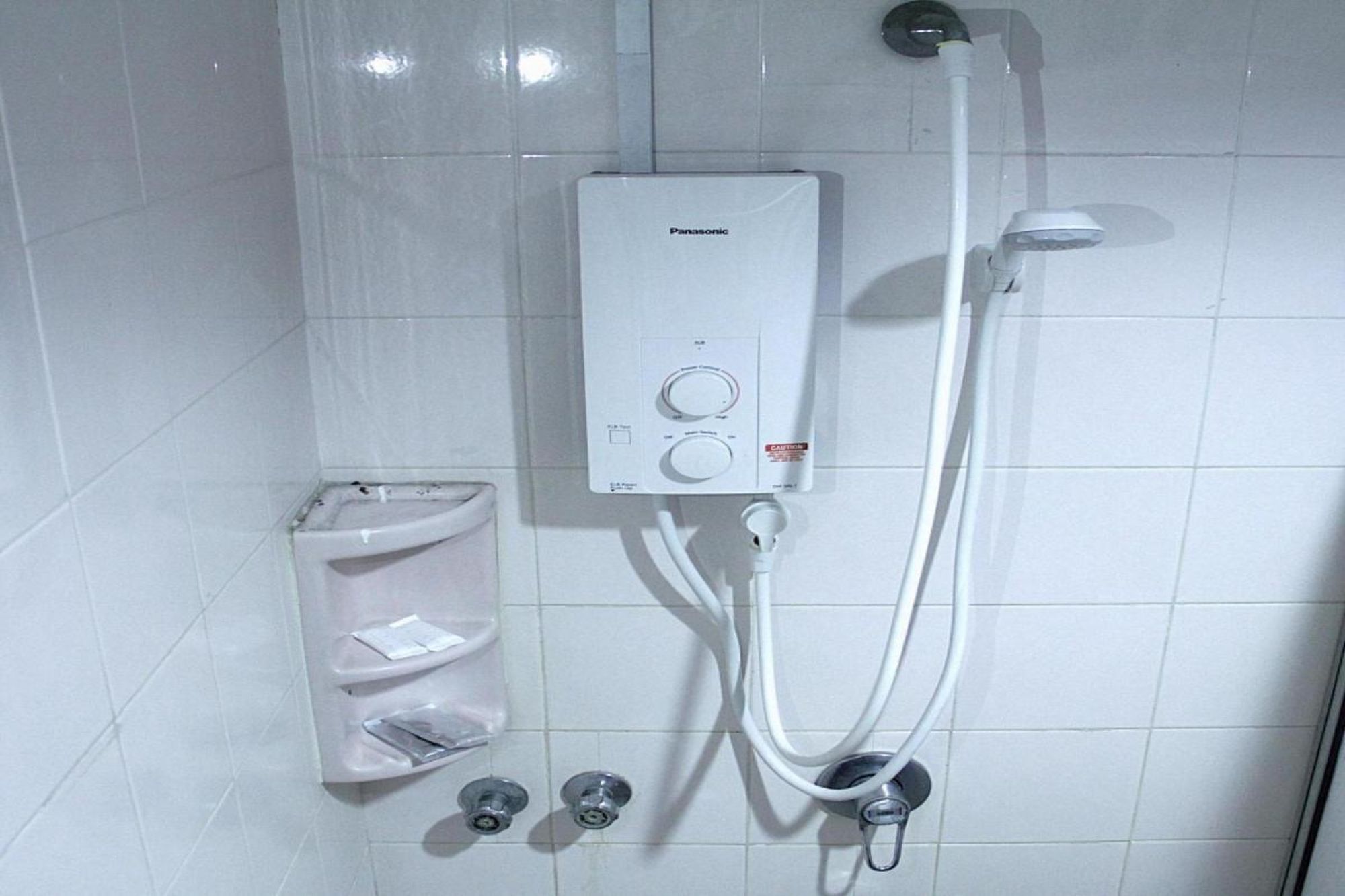 A bathroom with a white tiled wall, a Panasonic water heater, a handheld showerhead, and a soap dispenser.