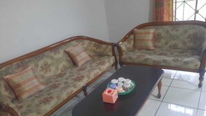 A living room with two floral-patterned sofas, a black coffee table with a pink object on it, and a window with curtains. - Hotel O Home Stay Syariah Annur (Jalaksana)