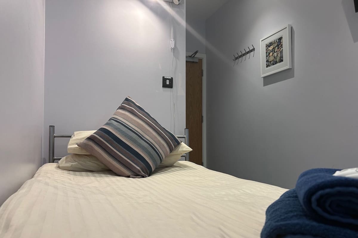 A neatly made bed with a striped pillow and rolled towels, a power outlet, a framed picture on the wall, and a door in the background.