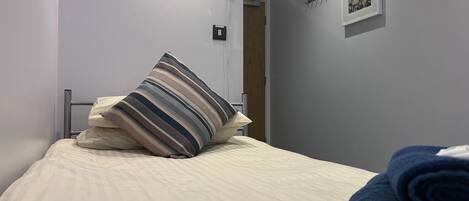Un lit bien fait, avec un oreiller à rayures et des serviettes enroulées, une prise de courant, un tableau encadré au mur et une porte visible au fond.