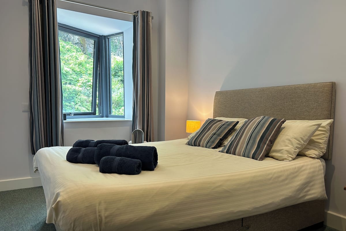 A neatly made bed with striped pillows and rolled towels, a bedside lamp, and a window with curtains.