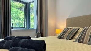 A hotel room with a bed, a window with curtains, a bedside lamp, and rolled towels on the bed. - Harbour Stays - The Marine Centre (Saundersfoot)