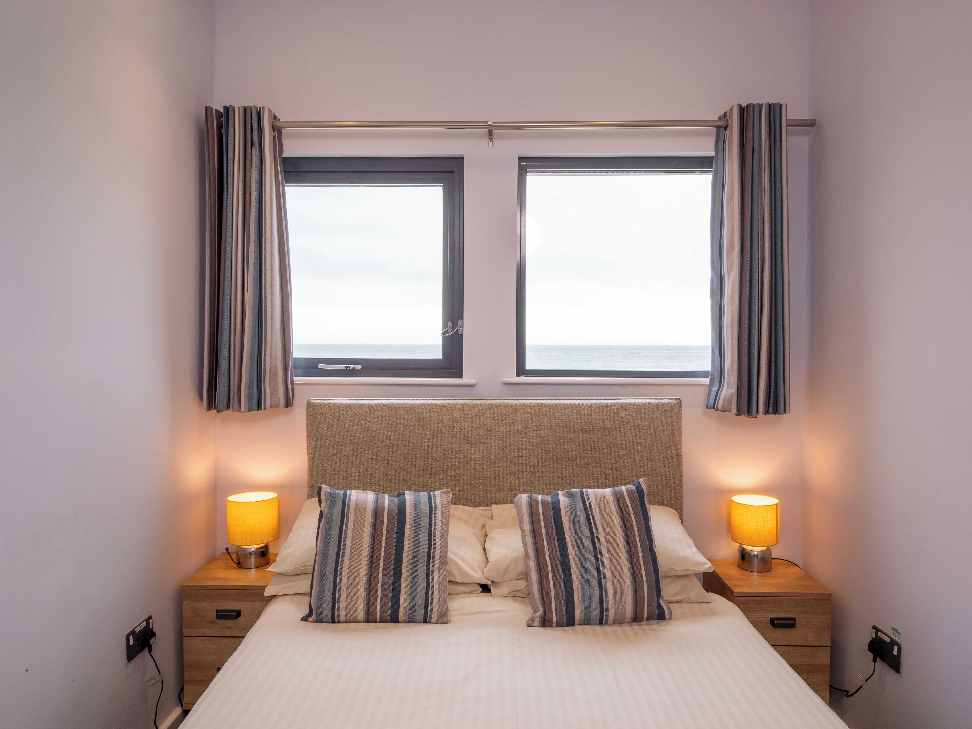 A hotel room with a bed, two bedside tables with lamps, a window with curtains, and a view of the sea.