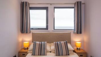 Ein Hotelzimmer mit einem Bett, zwei Nachttischen mit Lampen, einem Fenster mit Vorhängen und Blick auf das Meer.