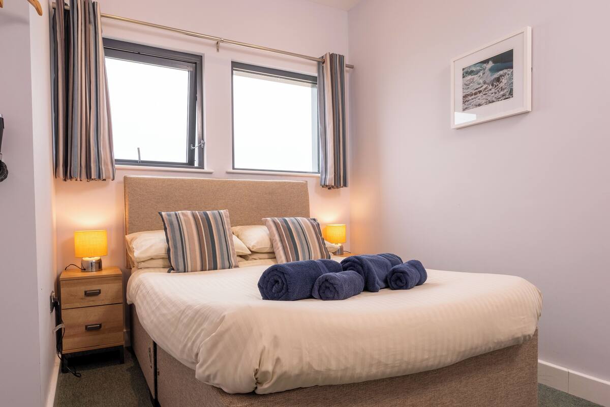 A neatly made bed with striped pillows, a bedside lamp, and rolled towels on a carpeted floor.