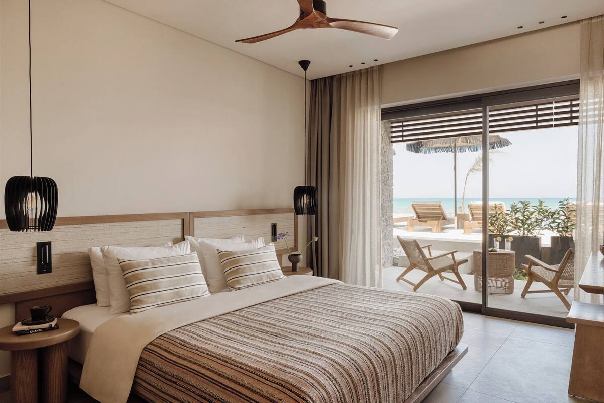Camera d'albergo con un letto grande, una ventola a soffitto, vista sulla spiaggia e un balcone con mobili da esterno.