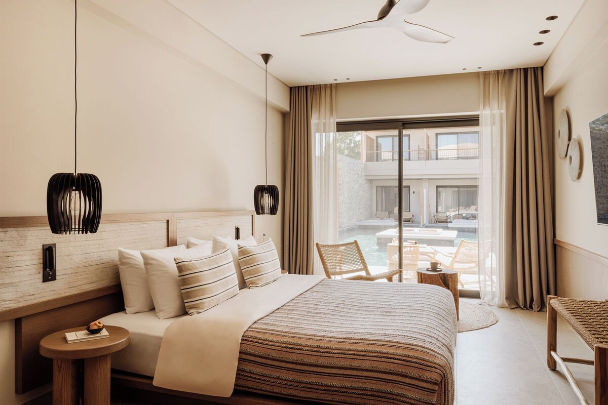 A modern hotel room with a large bed, a small table, a chair, and a view of a pool and outdoor area.