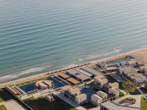 Vista aérea de un hotel frente al mar, con piscina y mobiliario en la playa, y una vista despejada del océano.