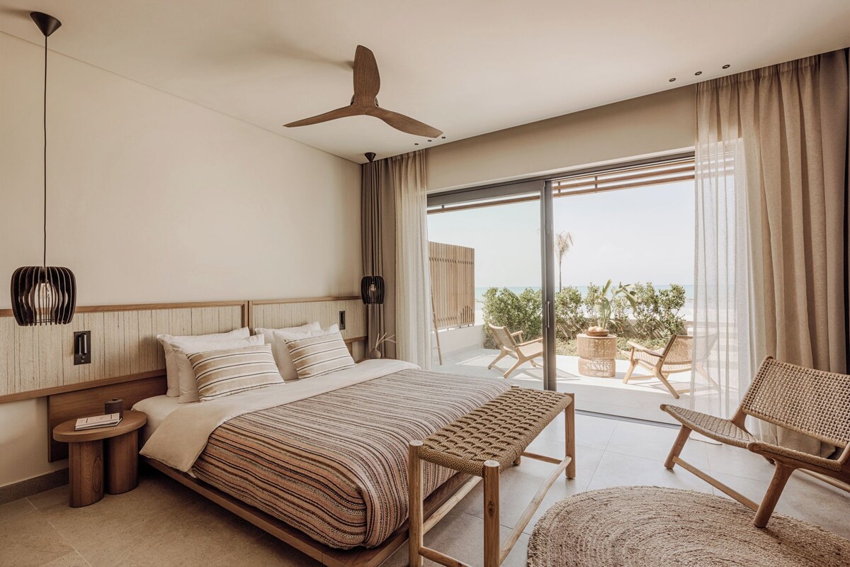 A modern bedroom with a large bed, a bench, a chair, and a view of the sea through a sliding glass door.