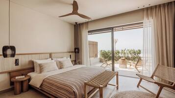 A modern bedroom with a large bed, a bench, a chair, and a view of the sea through a sliding glass door.