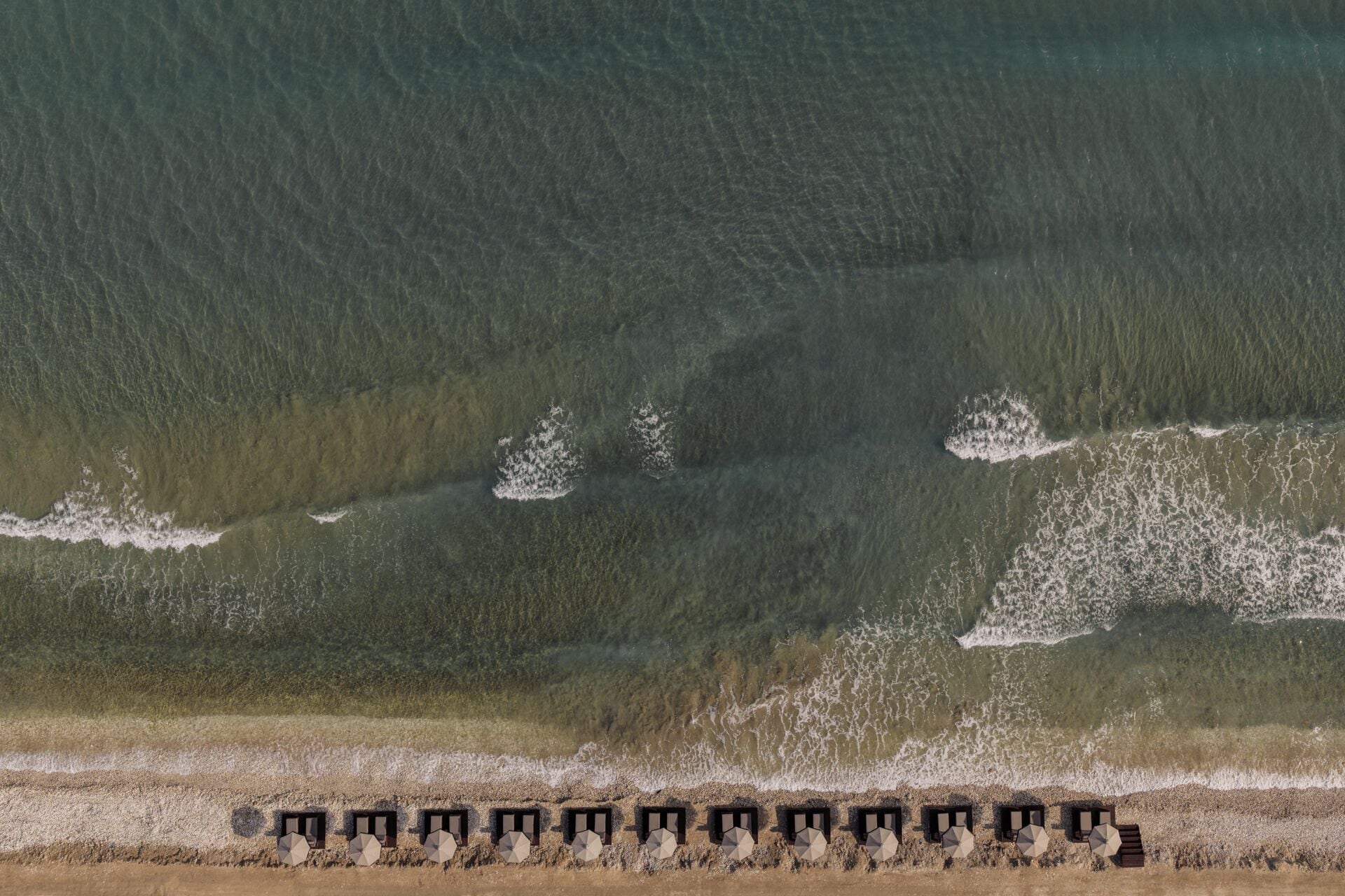 Vue aérienne d’une plage avec des vagues, une étendue de sable et une rangée de structures hexagonales surmontées de parasols.