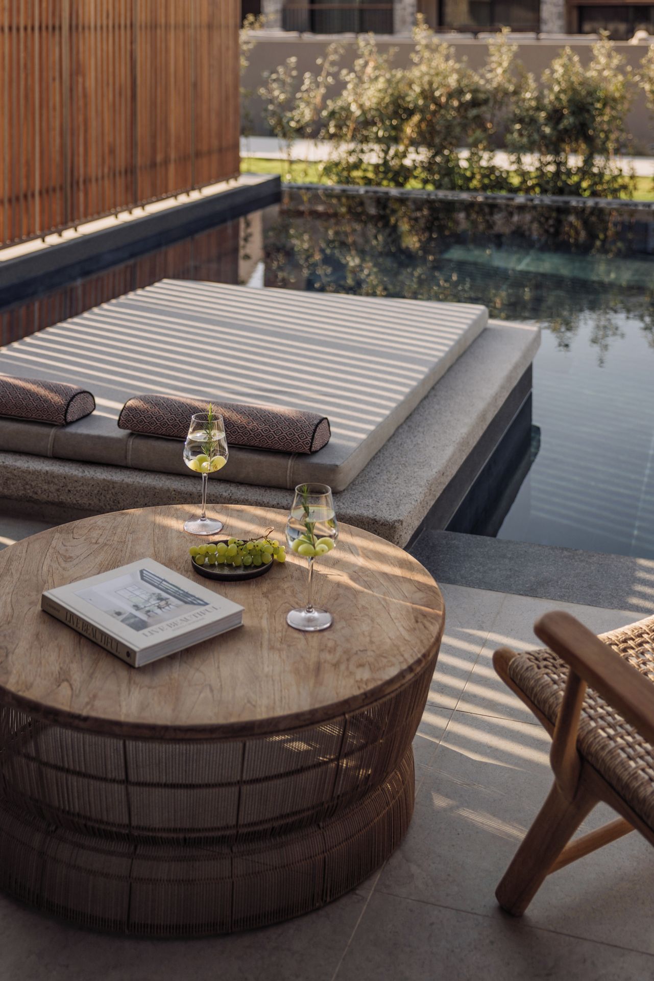 Una mesa de madera redonda con un libro, dos copas de vino y un bol de uvas en un patio con una piscina al fondo.