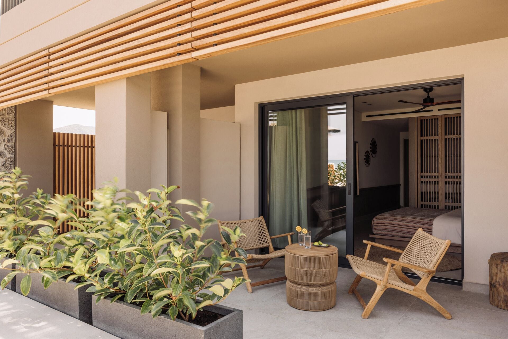 Un área de estar exterior moderna con mobiliario de mimbre, una mesa redonda de madera y una jardinera con follaje verde.