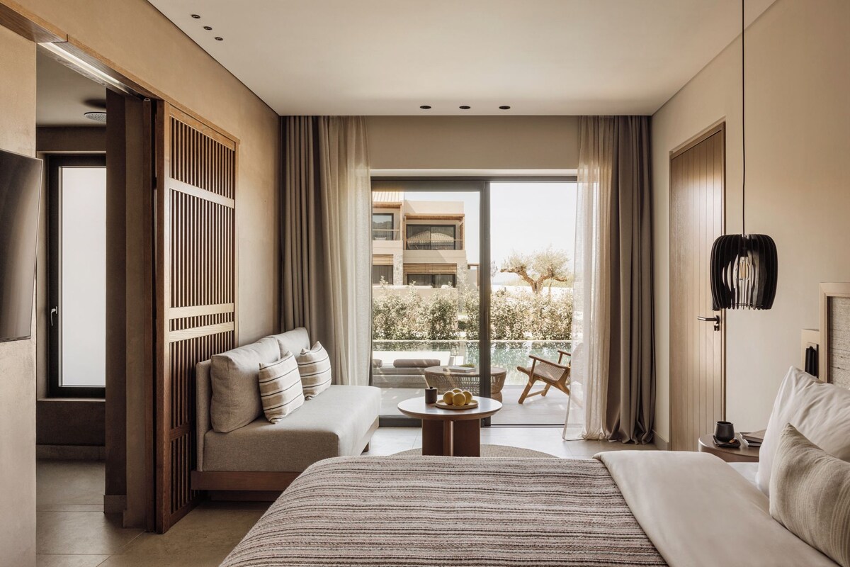 A modern hotel room with a large bed, a sofa, a small table, and a balcony with a view of a building and trees.