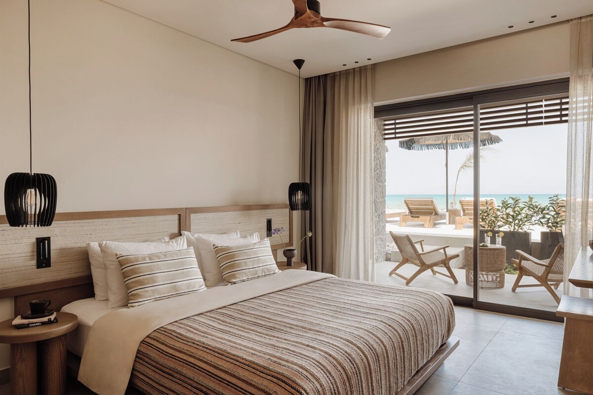 A hotel room with a large bed, a ceiling fan, a view of the beach, and a balcony with outdoor seating.