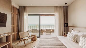 A hotel room with a bed, a desk, a chair, a small table, and a view of the ocean through a large window.