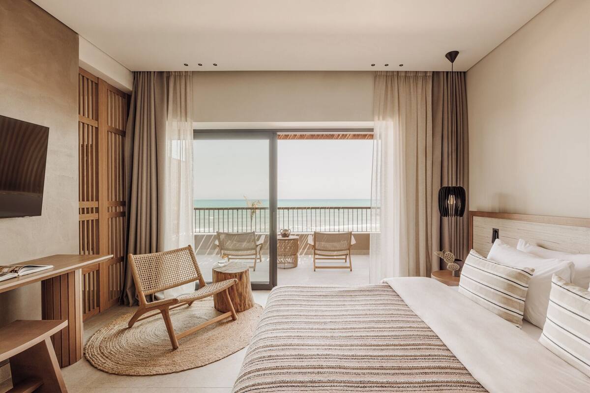 Una camera d'albergo con un letto, una scrivania, una sedia, un tavolino e vista sull'oceano attraverso una grande finestra.