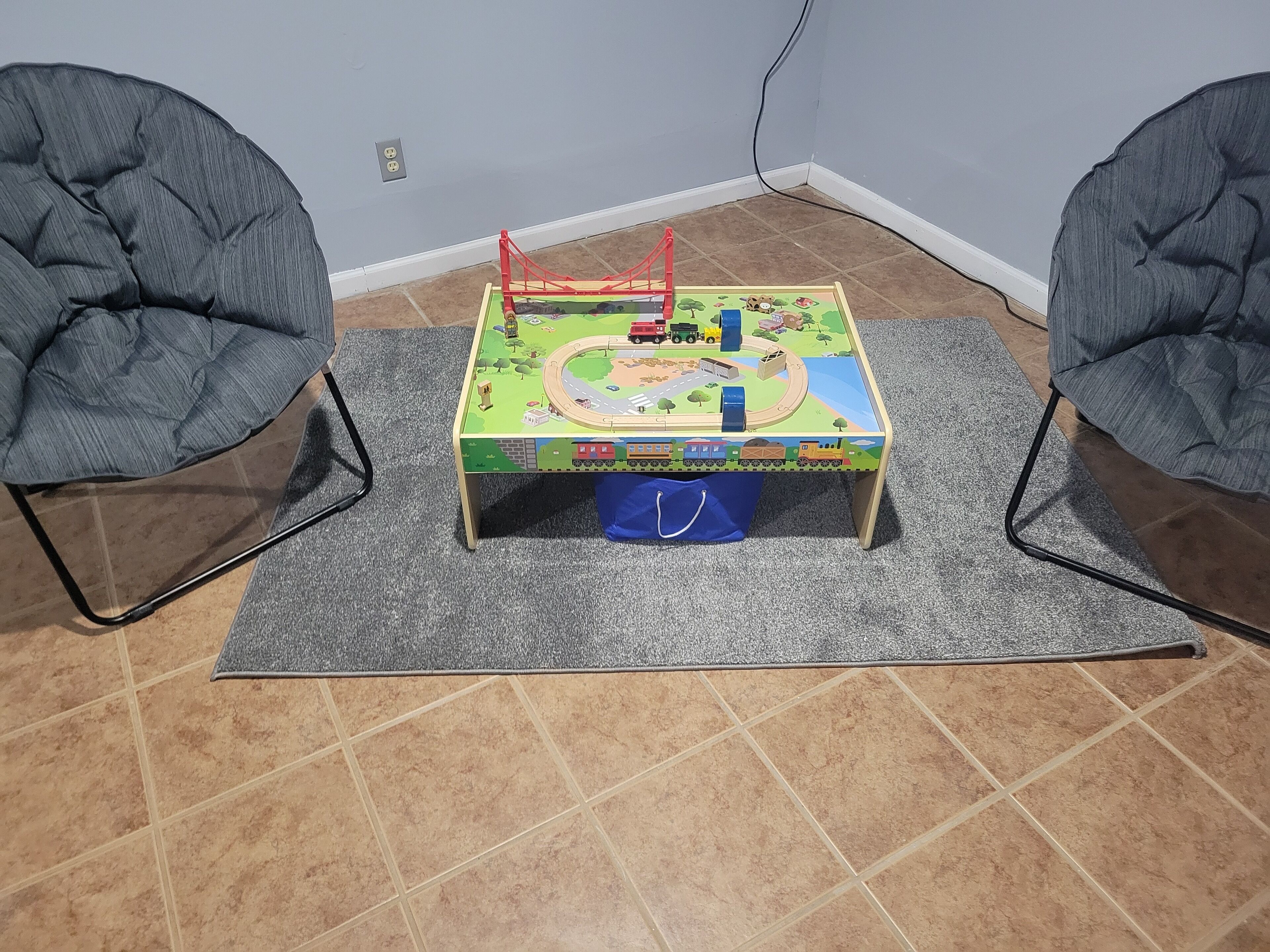 A small play area with a train set table, two chairs, and a grey rug on a tiled floor.