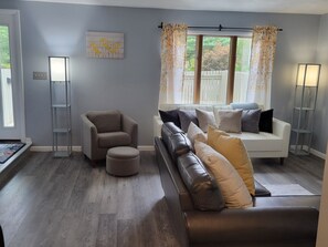 A modern living room with a grey sofa, a beige armchair, and a coffee table. There is a window with curtains, a floor lamp, and a painting on the wall. - Modern. 5 min to Penn State Breakfast included, King bed, Game Room! Toftrees (State College)