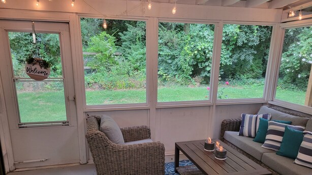 Una habitación acogedora con mobiliario de mimbre, un cartel que dice “Bienvenido” y ventanas grandes con vistas a un jardín.