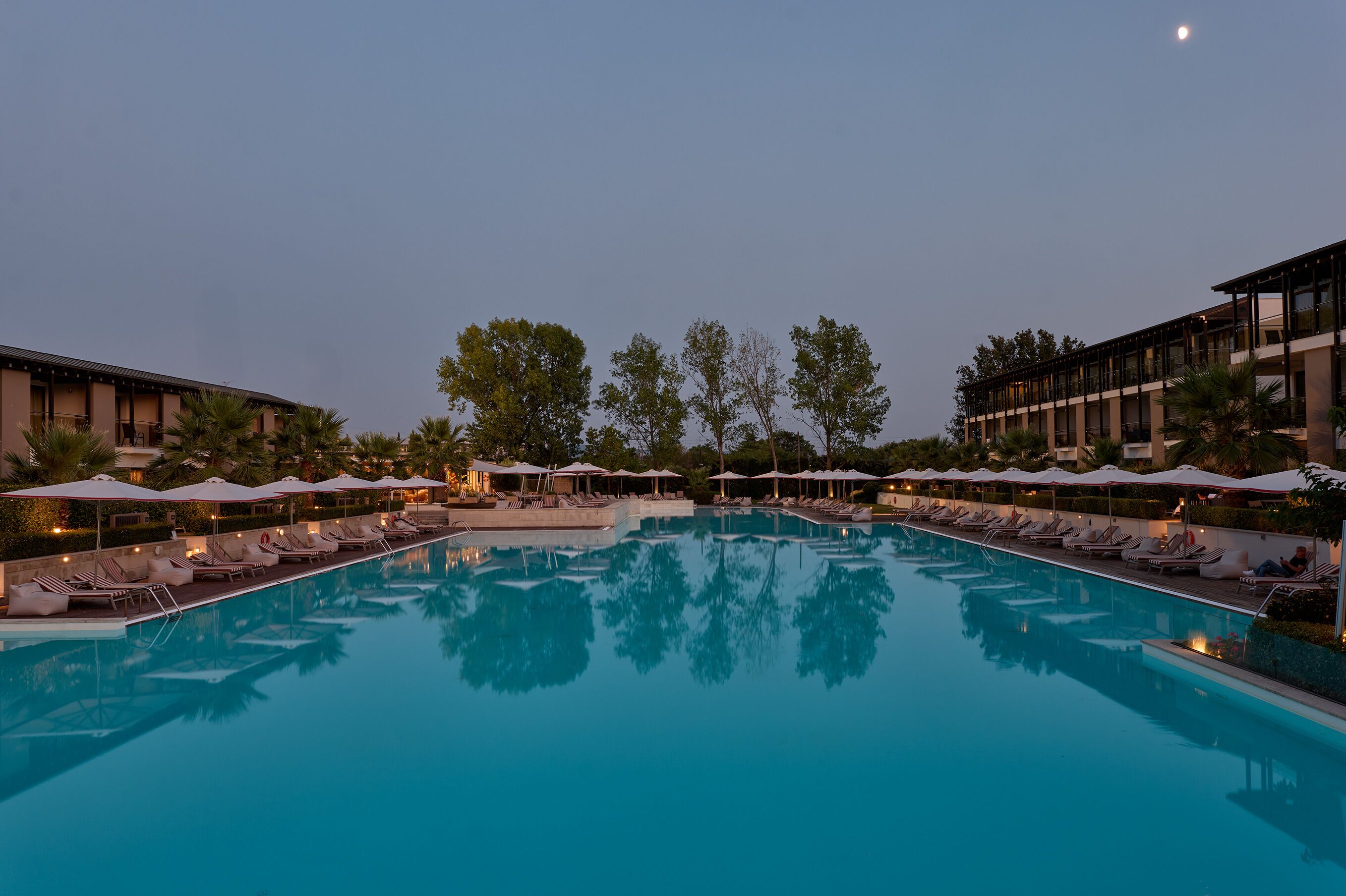 A large outdoor swimming pool with lounge chairs and umbrellas, surrounded by buildings and trees.