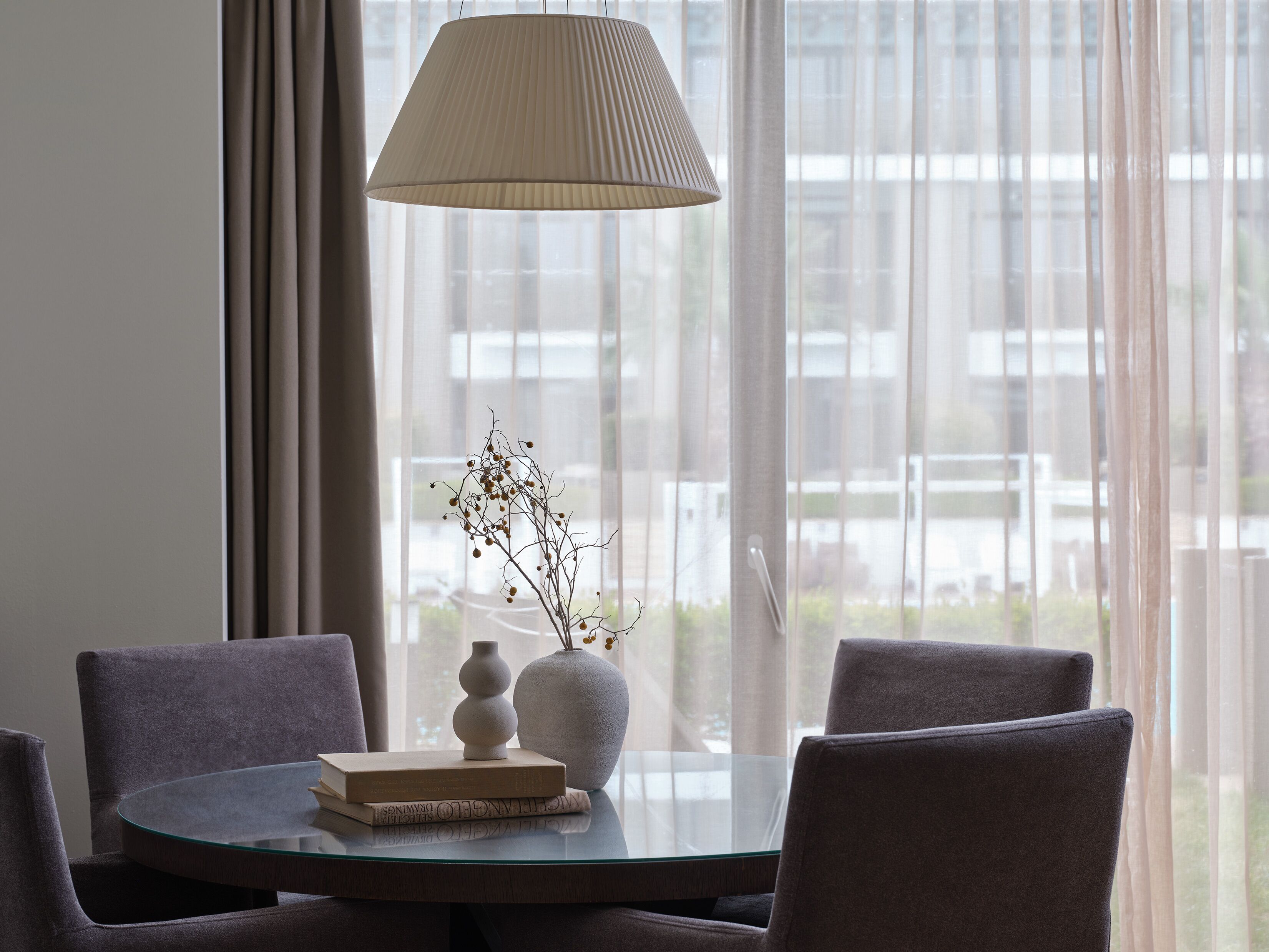 A modern dining area with a round glass table, two chairs, a vase with branches, and a lamp.