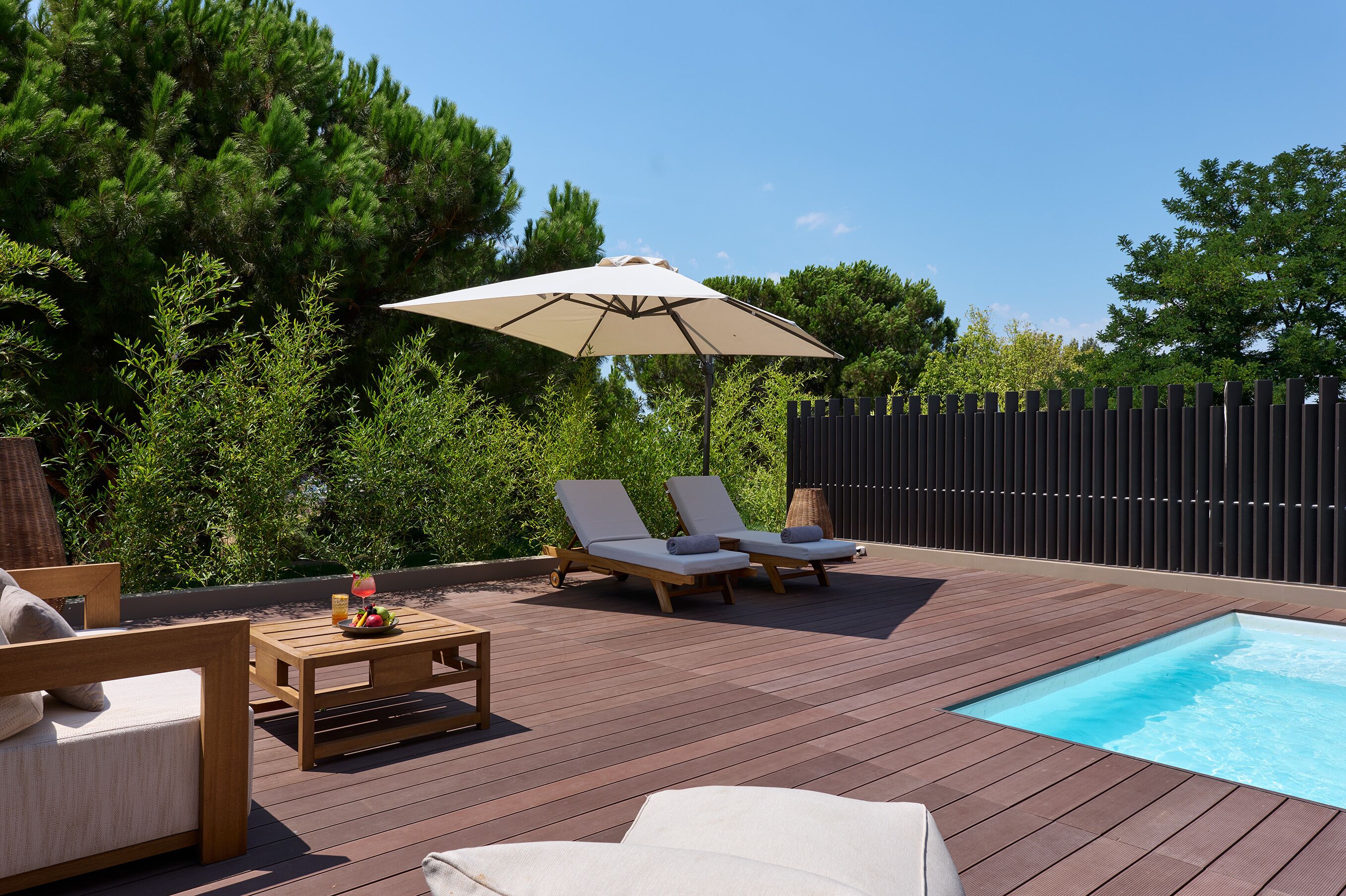 A wooden deck with two lounge chairs, a table with fruit and a drink, and a swimming pool.