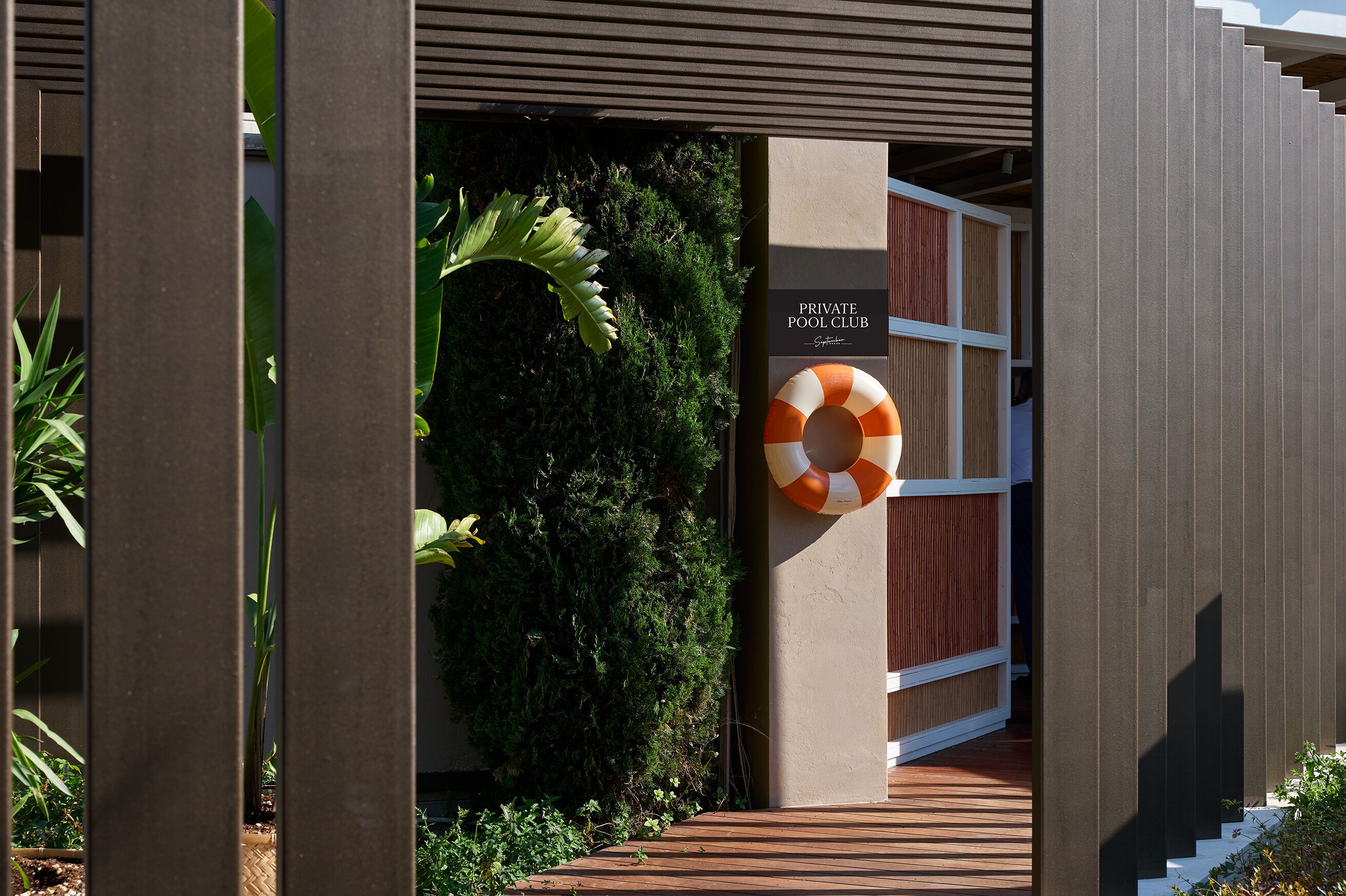 Entrada a un club privado con piscina, un muelle de madera, un flotador salvavidas y abundante vegetación.