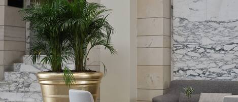 A modern lobby with a large potted plant, marble flooring, and a seating area with a sofa and armchairs.