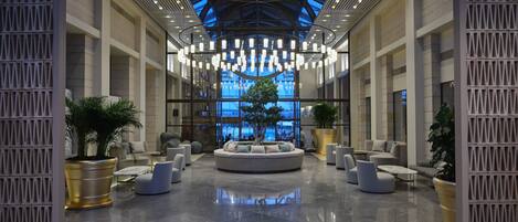 A spacious lobby with a central seating area, large potted plants, and a high ceiling with skylights.