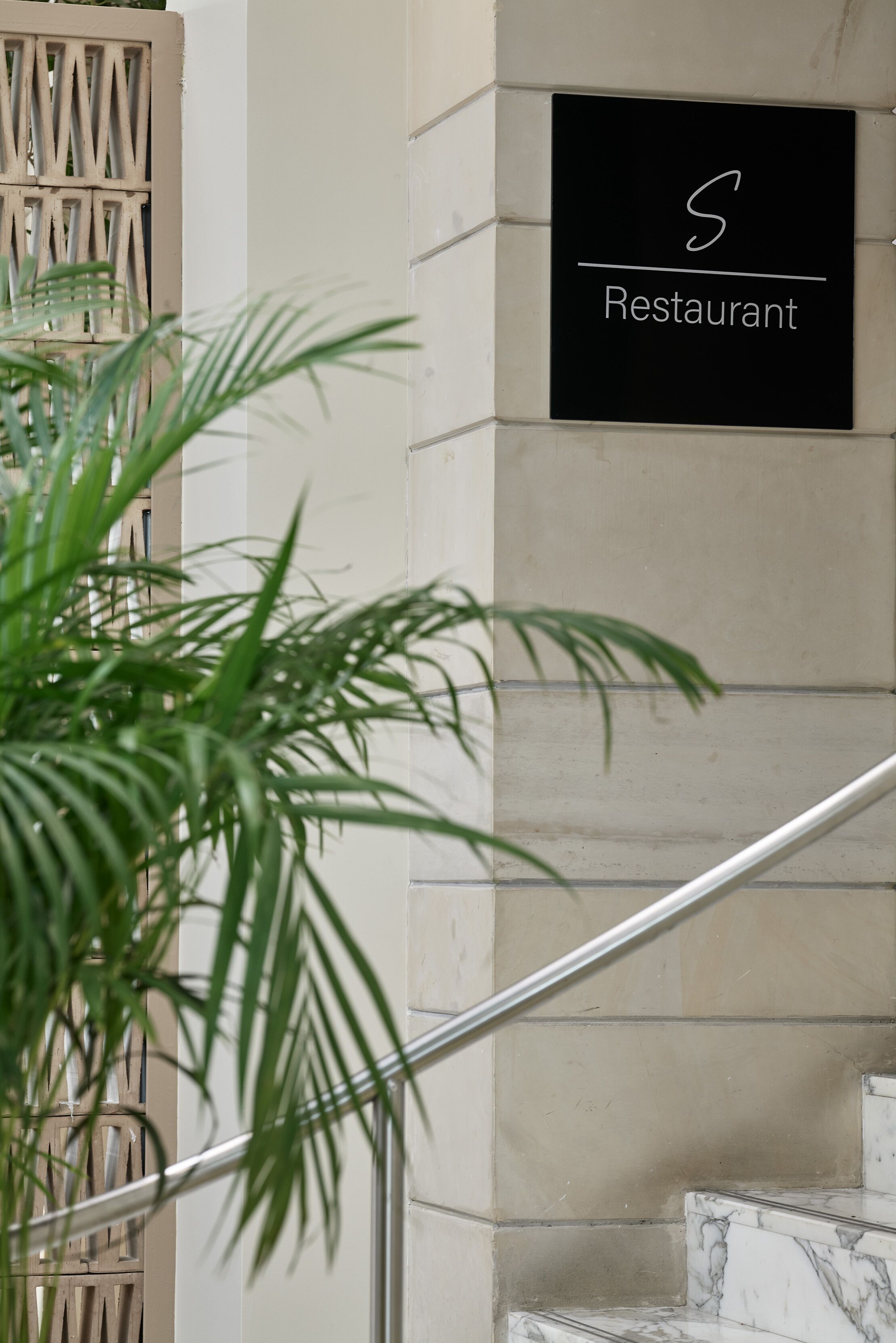 Un letrero de restaurante en la pared exterior de un edificio, una barandilla de metal y una planta en maceta.