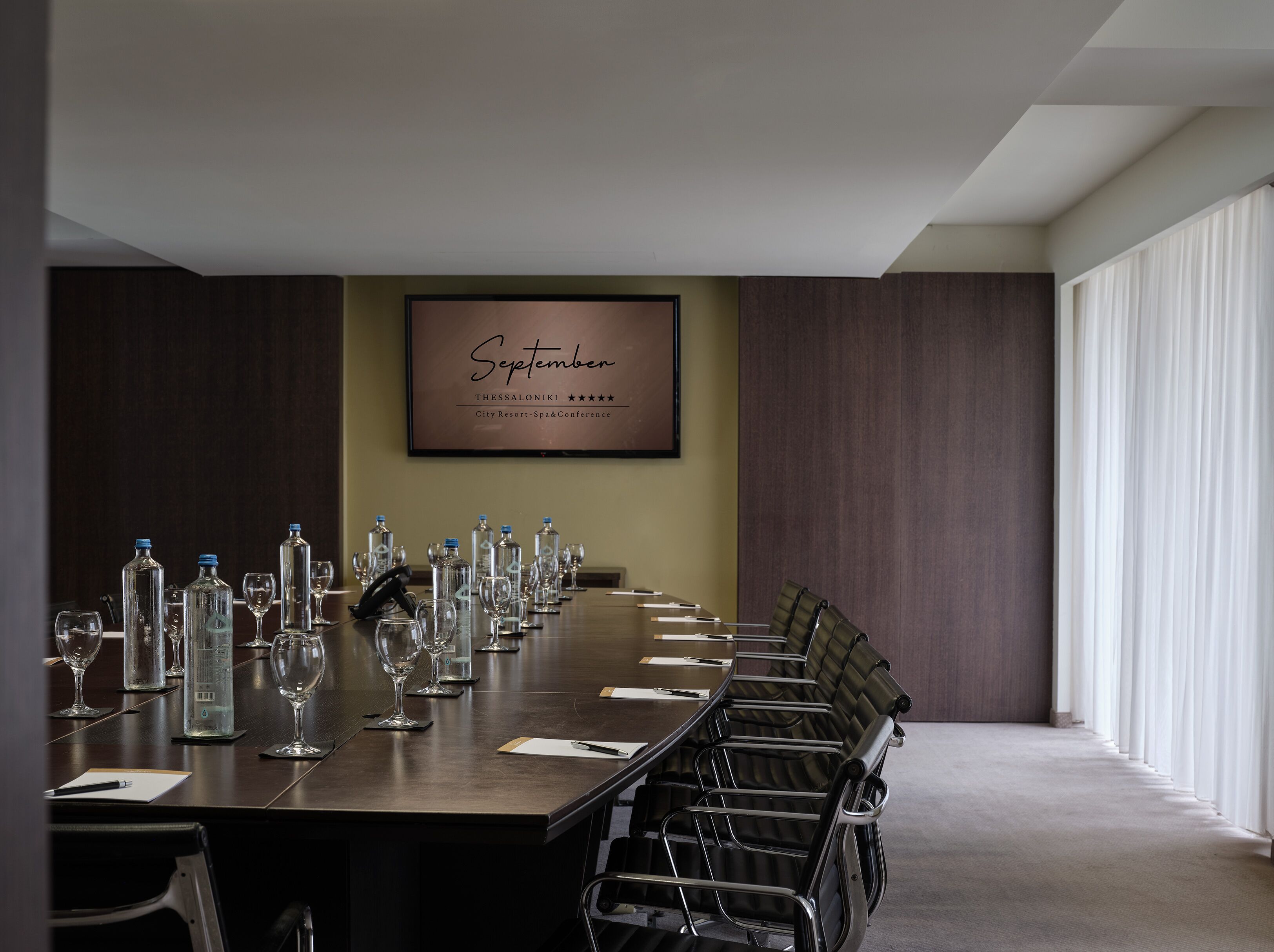 A conference room with a long table, chairs, and empty glassware. A framed sign on the wall reads "September Thessaloniki City Resort-Spa Conference".