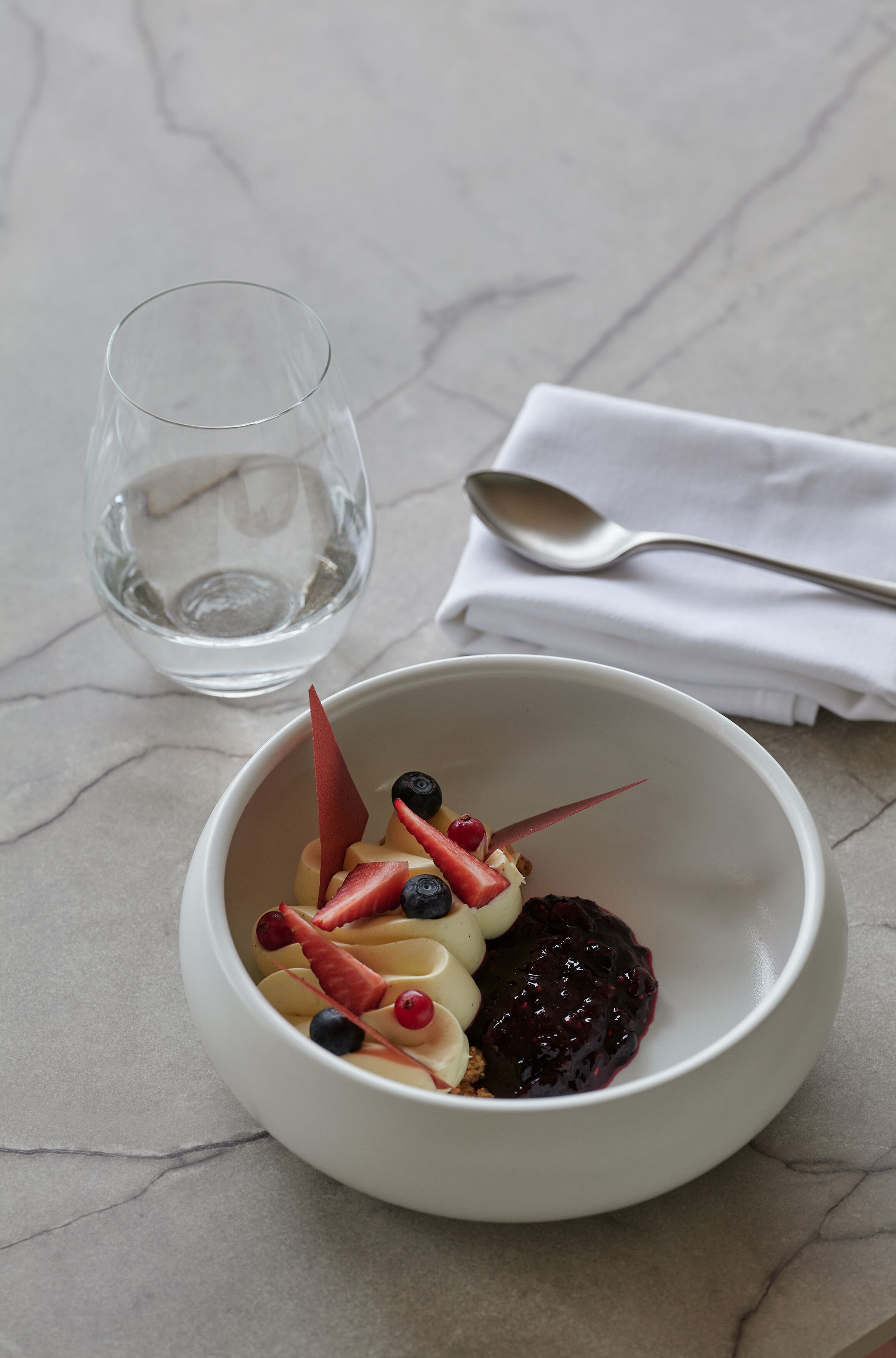 A white bowl with a dessert on a marble surface, a glass of water, and a spoon on a napkin.