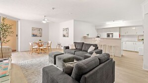 Een moderne woonkamer met een grijze hoekbank, een eetgedeelte met een houten tafel en stoelen, en een keuken met witte kasten en een magnetron.