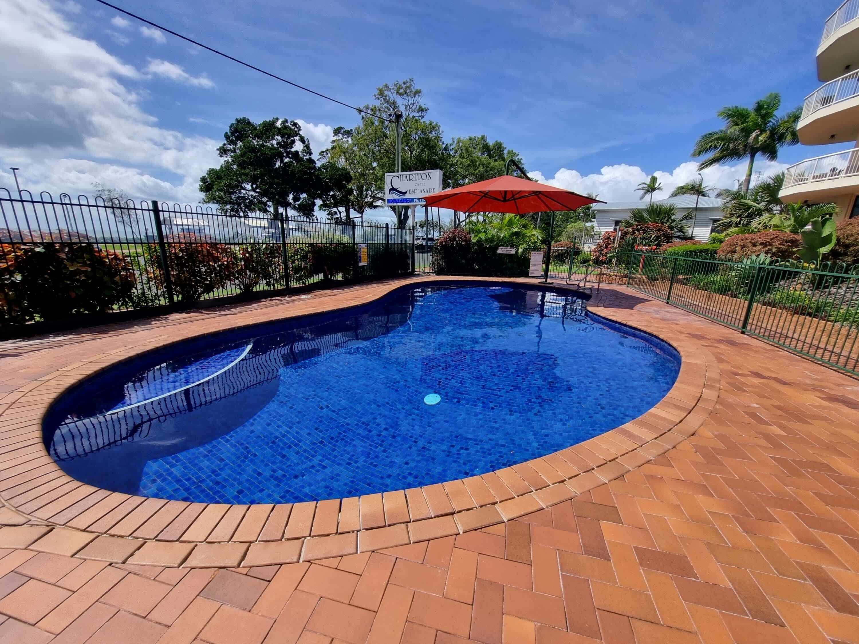 A swimming pool with a red umbrella, surrounded by a brick patio and a metal fence.