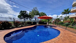 Una piscina con un paraguas rojo, rodeada por un patio de ladrillo y una valla de metal.