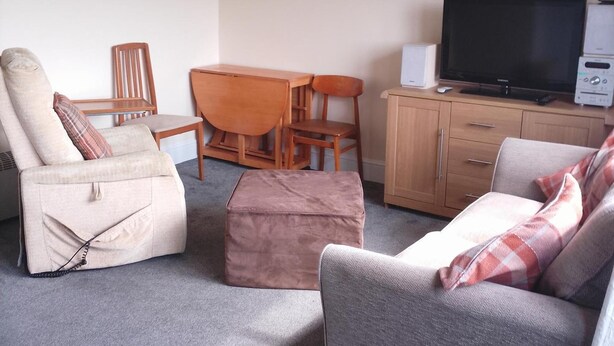 Ein Wohnzimmer mit Sofa, Sessel, Hocker, Holztisch, Stühlen, einem Fernseher und einem Schrank.