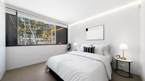 Un dormitorio moderno con un ventanal grande, una cama con ropa de cama blanca, una mesita de noche con una lámpara y un pequeño reloj.
