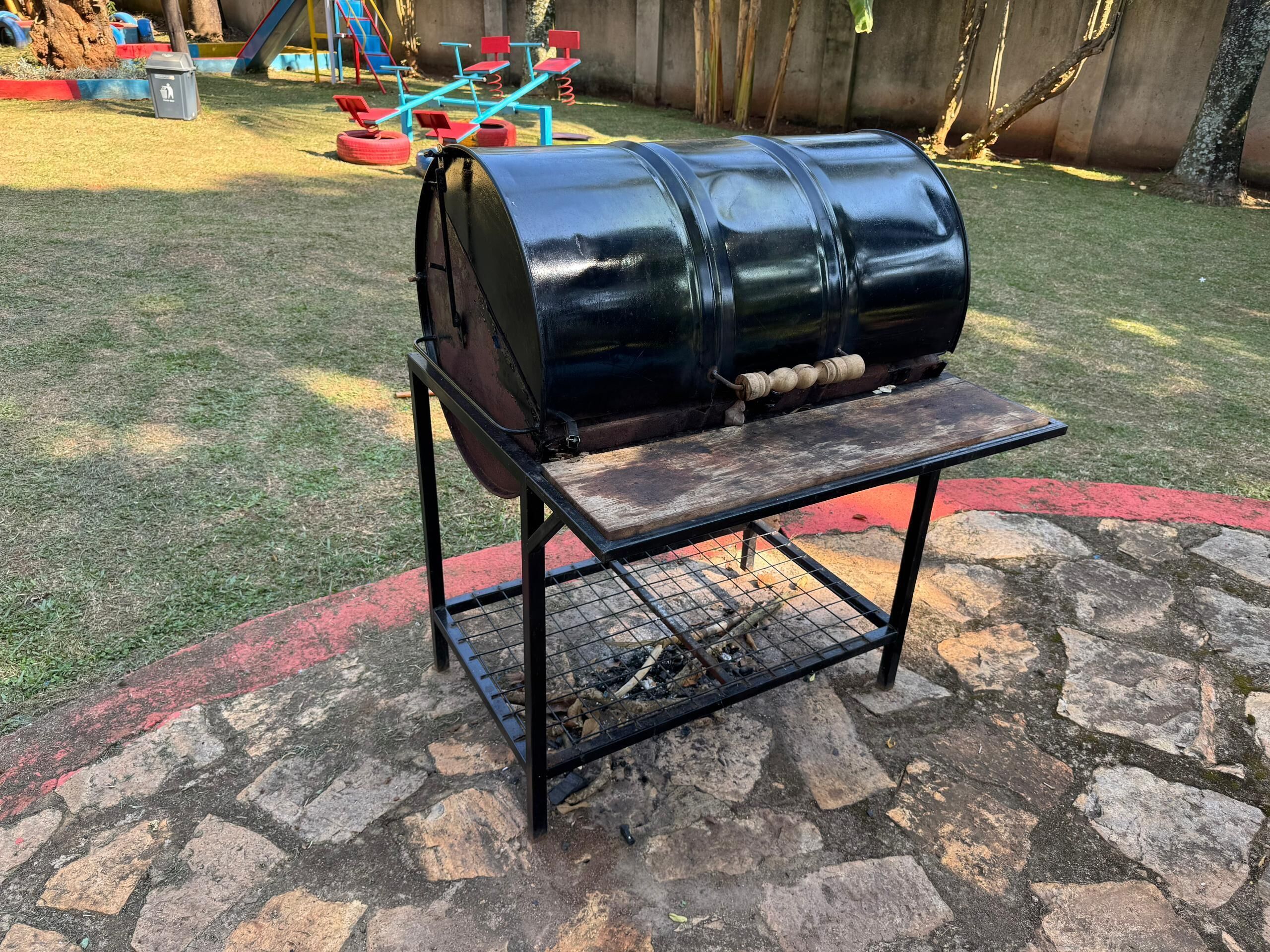 Una barbacoa amb estructura metàl·lica i prestatge de fusta, situada en una zona pavimentada de pedra, amb gespa al fons.