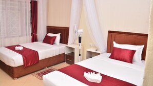 Habitación de hotel con dos camas, cada una con almohada roja y blanca, una mesita de noche con lámpara y un dosel blanco sobre las camas.
