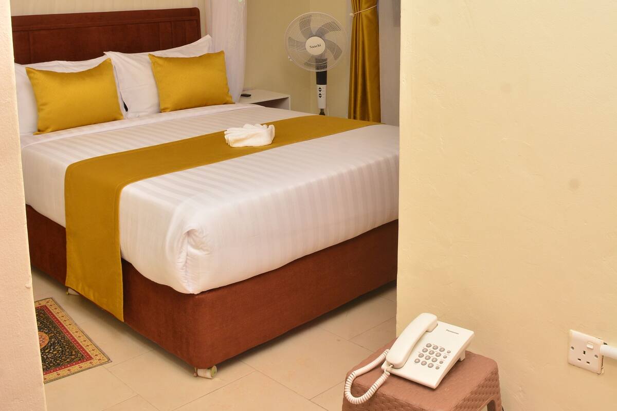A hotel room with a bed, a bedside table, a fan, and a telephone on a stool.