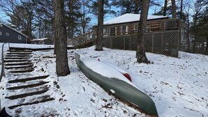Une structure en bois recouverte de neige, avec un canoë à proximité, entourée d’arbres et avec une maison en arrière-plan.
