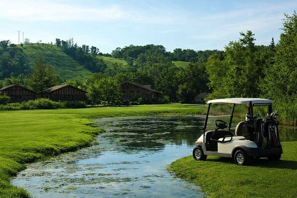 A golf cart parked by a calm river, surrounded by lush greenery and wooden buildings. - Morning Tee Times  Walk to Golf  Summer Stay (Mancelona)