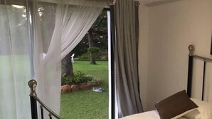 A bedroom with a bed, a window with curtains, and a view of a garden. - 14 on Carnarvon (Bulawayo)