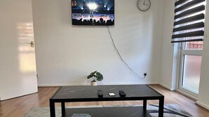 A room with a television mounted on the wall, a clock, a small potted plant, a coffee table with remotes, and a window with blinds. - Hive Stays Close to Abbeywood Free Parking Wifi (Erith)