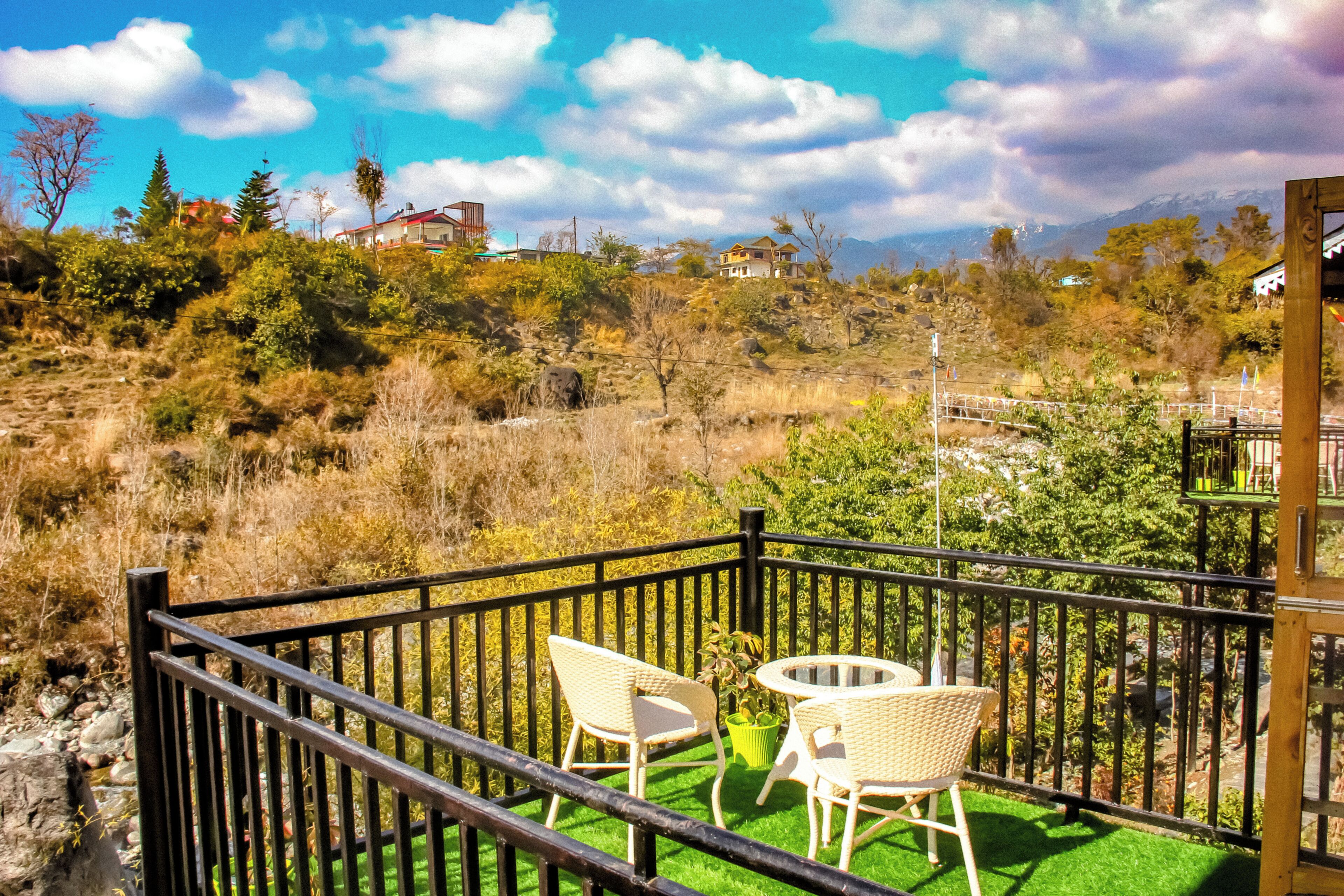Un balcón con dos sillones de mimbre y una pequeña mesa redonda, con vistas a un paisaje montañoso de vegetación escasa y algunos edificios a lo lejos.