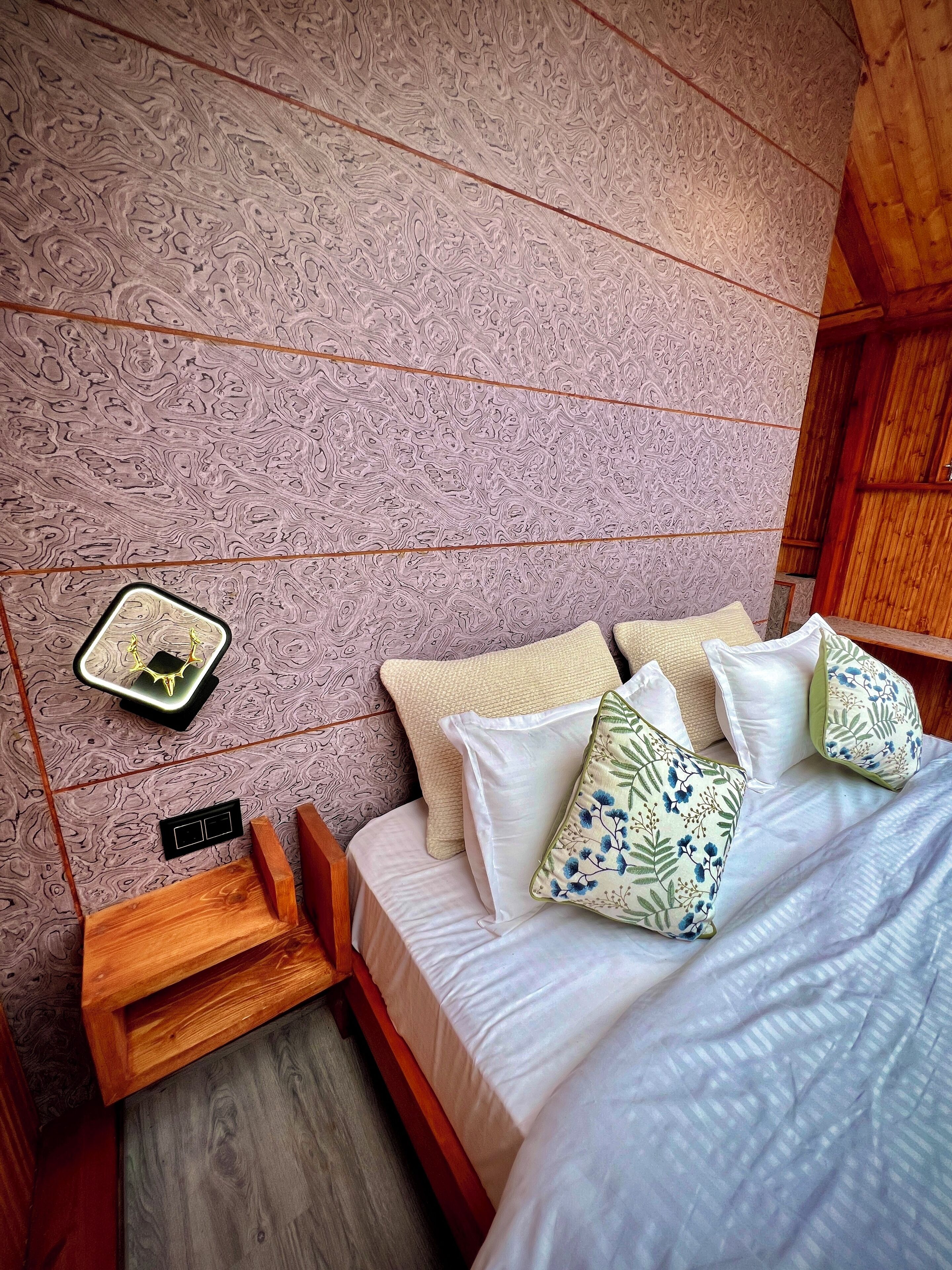 Un dormitorio con cama, almohadas, una mesita de noche y una lámpara de pared.