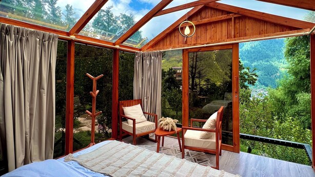 Ein Schlafzimmer mit einem großen Bett, zwei Holzsesseln und Blick auf einen Wald durch eine Glasdecke.