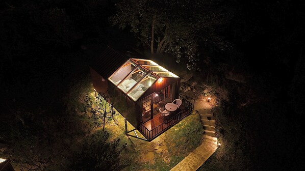 Une petite cabane illuminée, dotée d’un étage supérieur vitré, d’une terrasse en bois et d’un escalier en pierre menant à l’entrée.